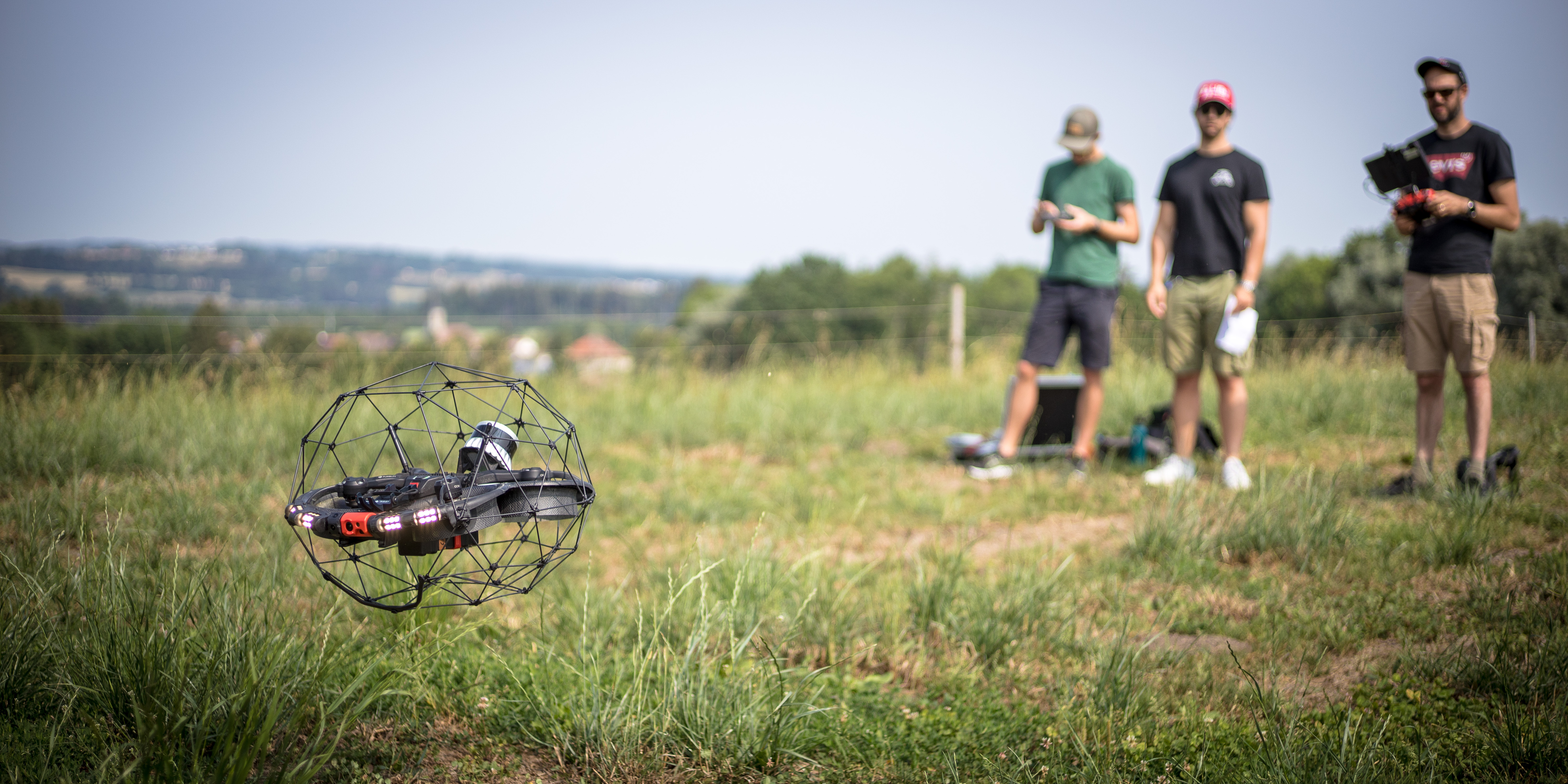 The Elios 3 drone taking off in a field of grass, with 3 people blurred out of focus behind the drone